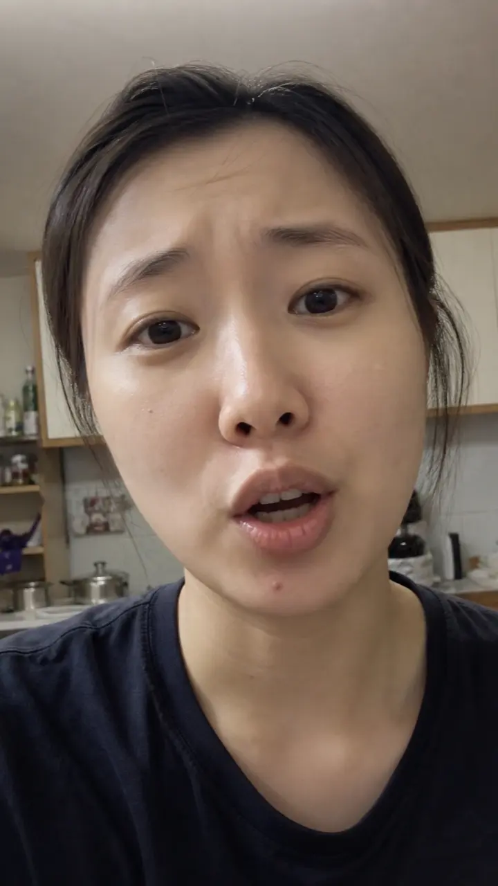 A woman talking to a phone camera in a small kitchen under flat overhead light.