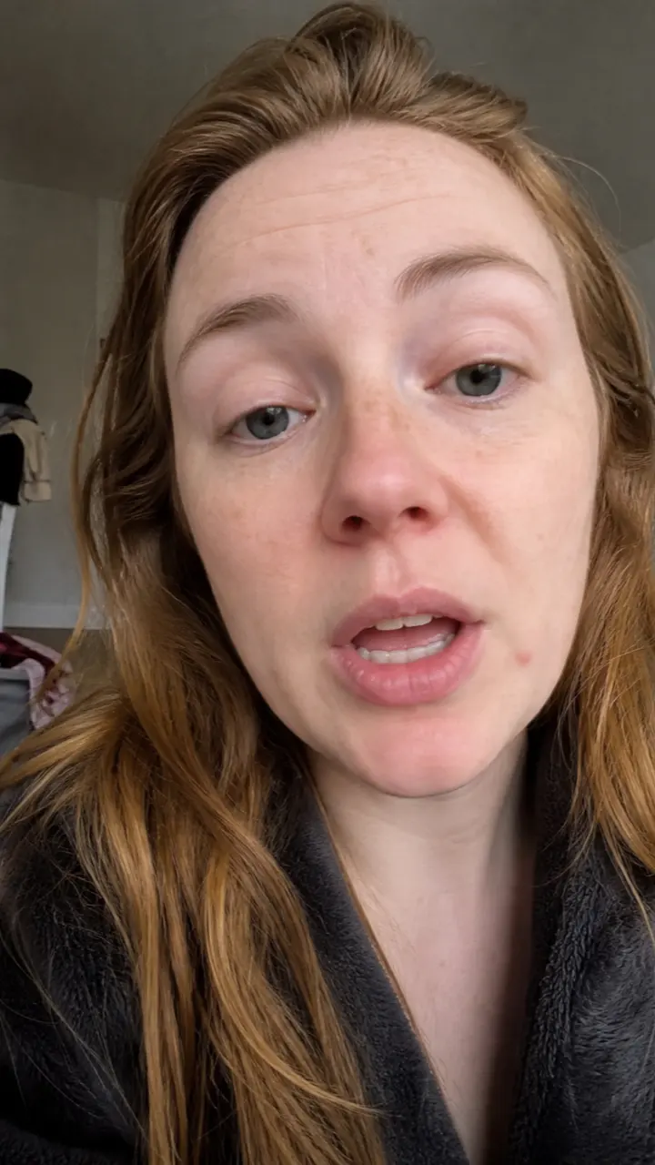 A woman talking to a phone camera in a cluttered bedroom under flat grey daylight.