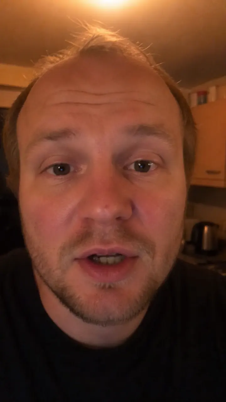 A man talking to a phone camera in a kitchen at night under a single warm bulb.