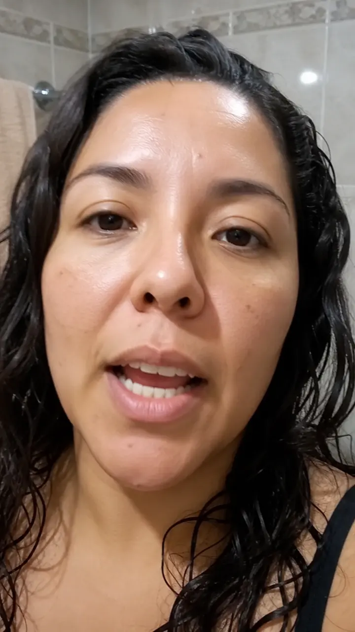 A woman with wet hair talking to a phone camera in a tiled bathroom.