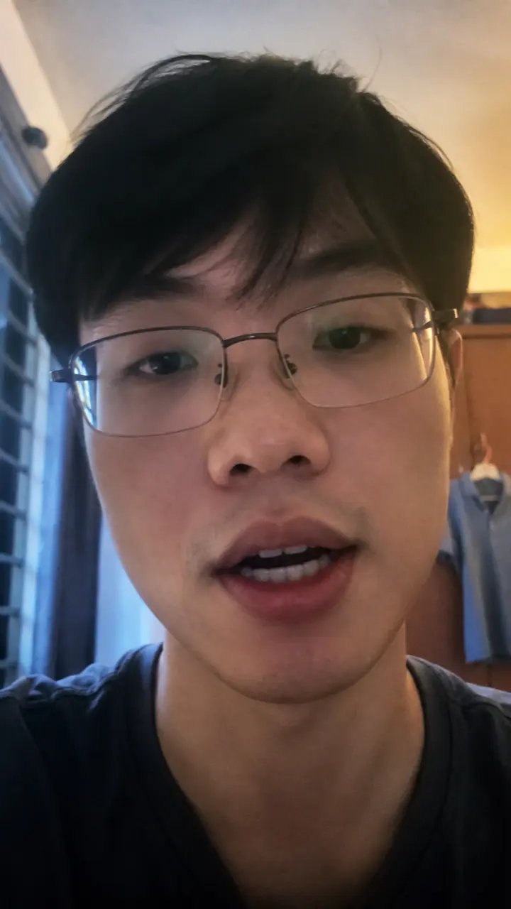 A man in glasses talking to a phone camera in a small room, lit from a window on one side.
